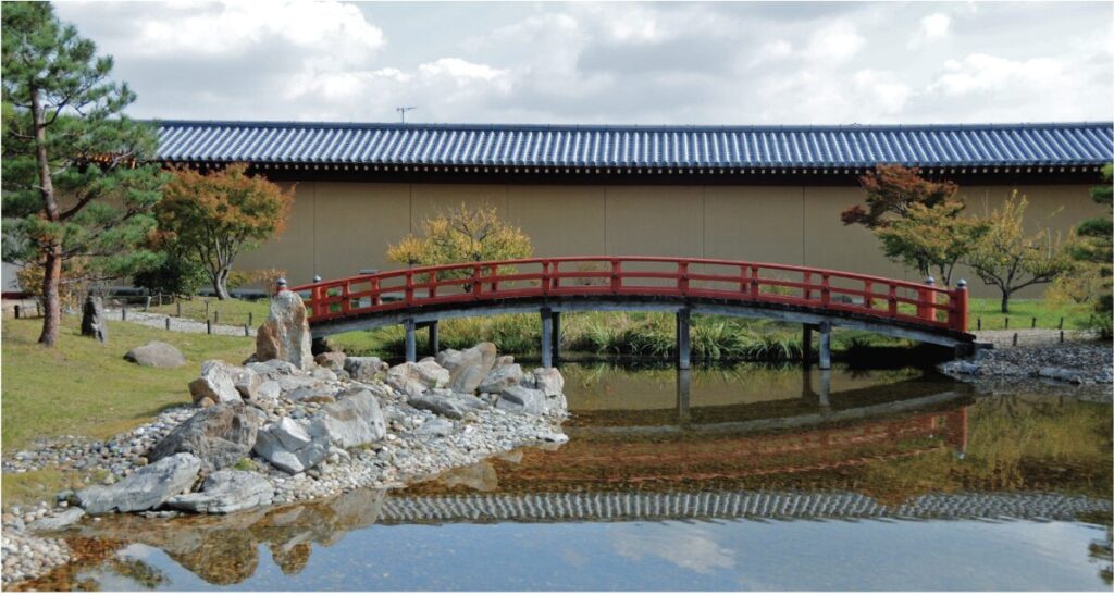 A red arched bridge over a Japanese garden pond with a rammed earth wall behind