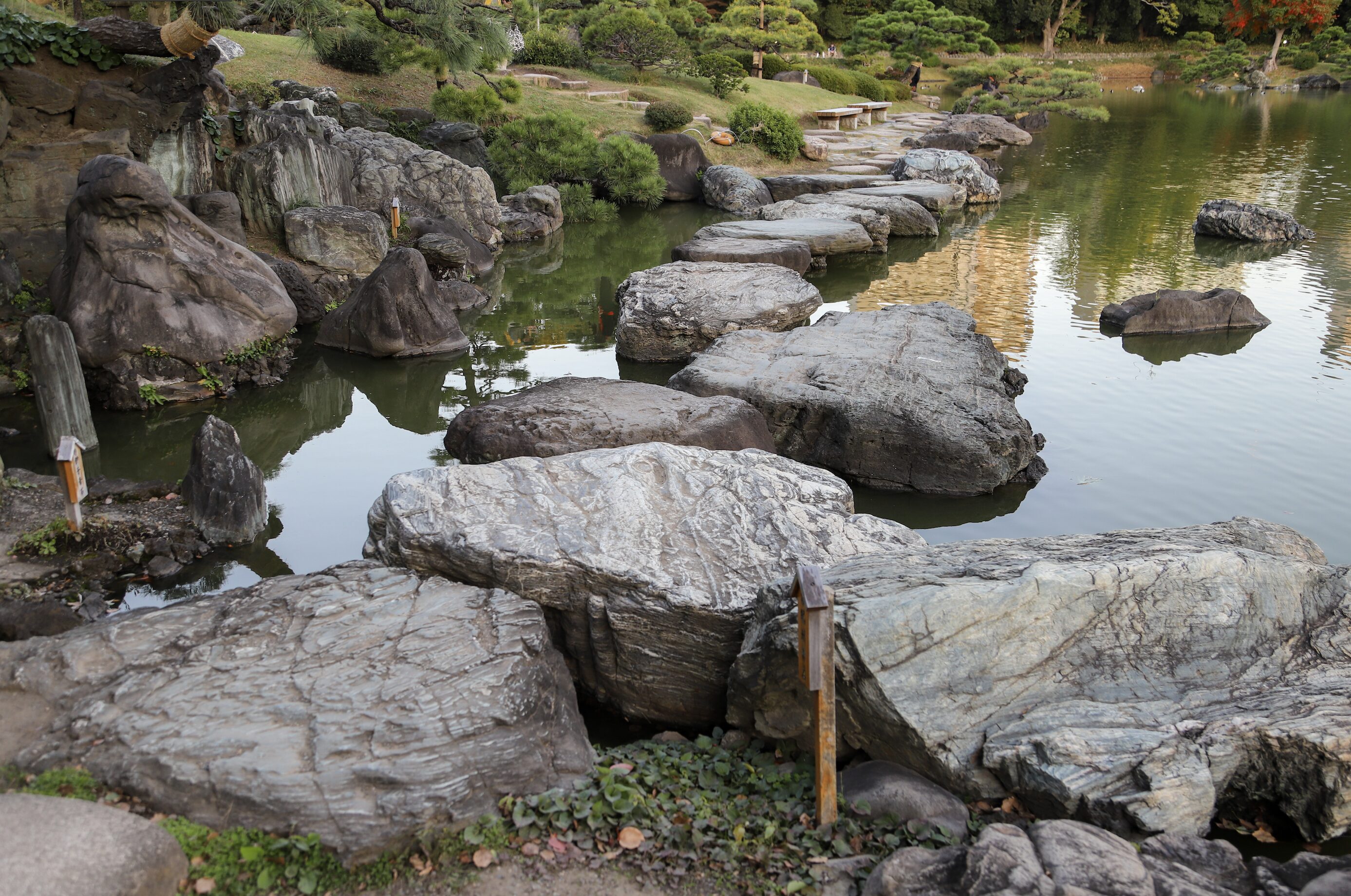 7. Kiyosumi- amzing rocks rocks A Japanese garden scene featuring a series of large, irregularly shaped stepping stones forming a pathway across a calm pond. The water reflects the surrounding lush greenery, manicured shrubs, and trees. In the background, a stone pathway, wooden benches, and additional rocks add to the serene landscape.