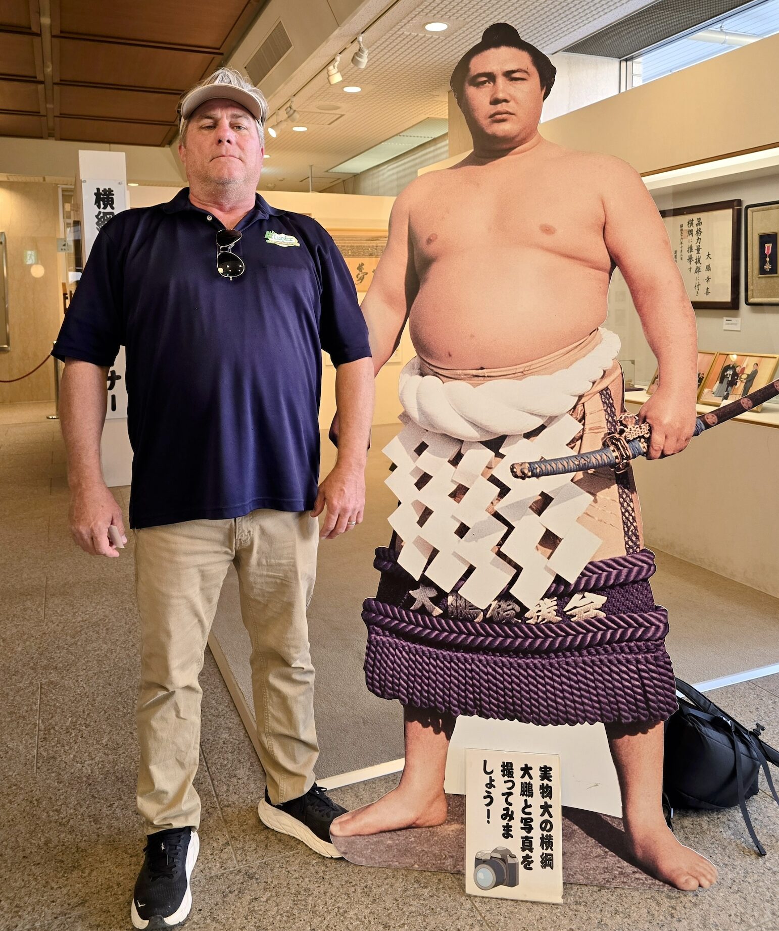 9. Tom with Sumo cut-out A man wearing a navy blue polo shirt, beige pants, black sneakers, and a beige cap stands next to a life-size cutout of a sumo wrestler inside a museum or exhibition space. He has sunglasses hanging from his shirt collar and a neutral expression. The sumo wrestler cutout depicts a traditional yokozuna (grand champion) with a thick ceremonial rope (shimenawa) around his waist, holding a sword. A sign at the bottom of the cutout contains Japanese text and an icon of a camera, suggesting visitors take a photo. The background features framed pictures and artifacts on display in a well-lit room.